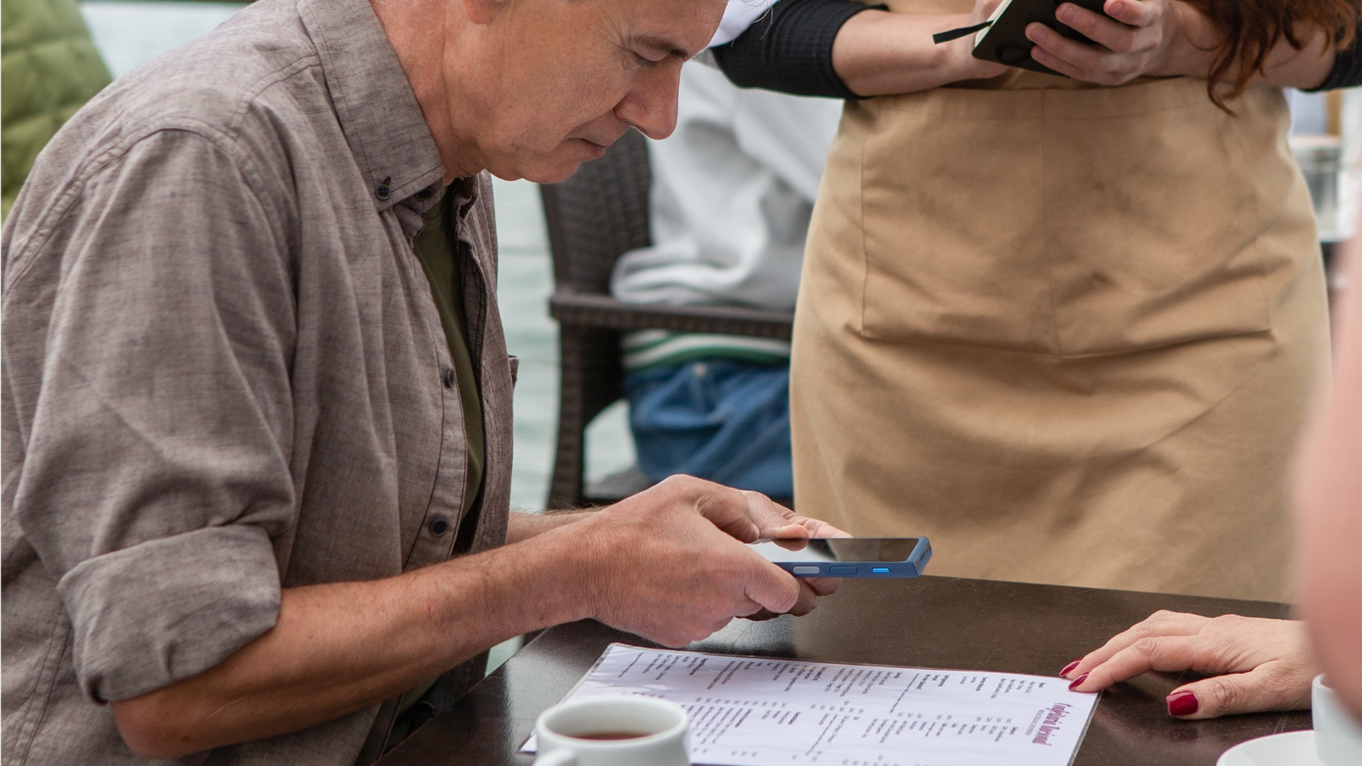 Un homme utilise son traducteur Vasco pour comprendre le menu d'un restaurant pendant que la serveuse attend la commande.