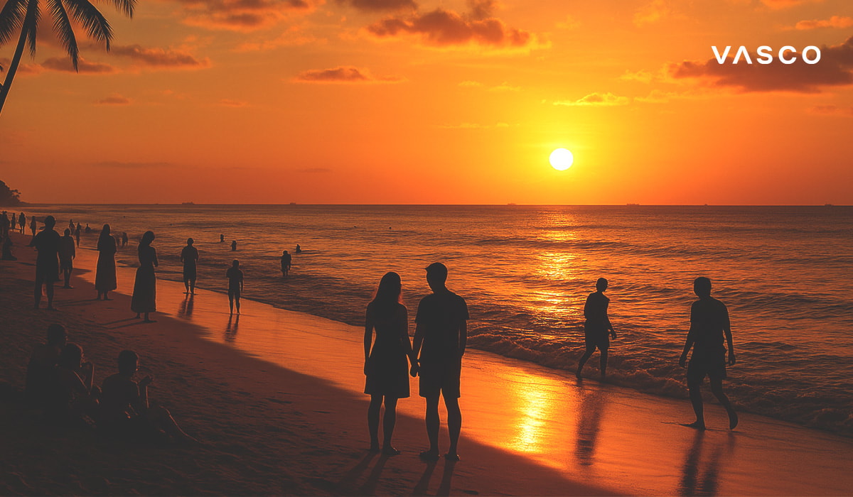 Un couple admire un magnifique coucher de soleil orangé sur une plage de Bali, entouré des silhouettes d'autres personnes dans la lumière déclinante.