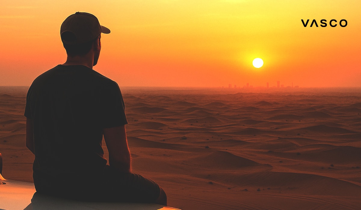 Un homme est assis de dos dans le désert au coucher du soleil, contemplant au loin un horizon de dunes et la silhouette d'une ville, avec le logo de Vasco en surimpression.