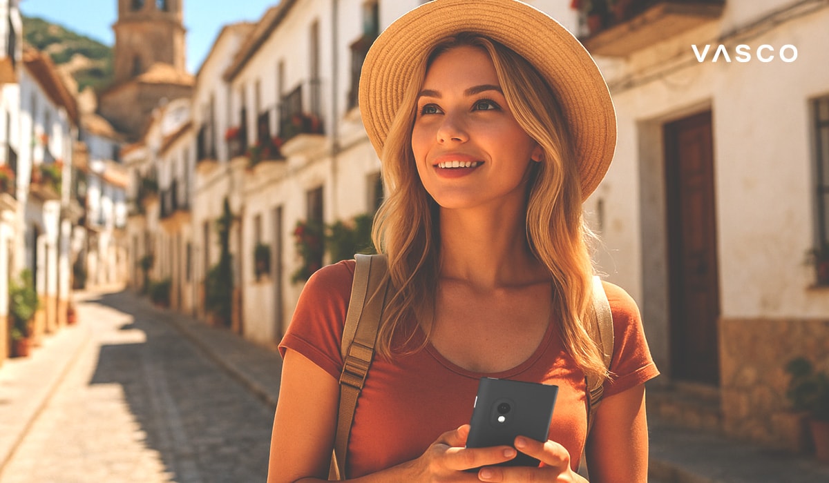 Une jeune femme blonde souriante, portant un chapeau de paille et un sac à dos, utilise son téléphone portable en marchant dans une rue pavée en Espagne.