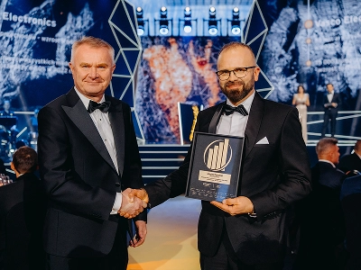 Plan moyen-long montrant deux hommes en tenue de soirée qui se serrent la main. L'homme de droite, Maciej Góralski, tient un trophée encadré orné d'un emblème doré, indiquant qu'il s'agit du prix de l'« Entrepreneur de l'année 2022 ». À l'arrière-plan, flou, une scène présente des structures géométriques abstraites et des motifs graphiques dynamiques bleus et rouge-orange. Plusieurs invités sont assis à des tables dans la salle de réception faiblement éclairée.