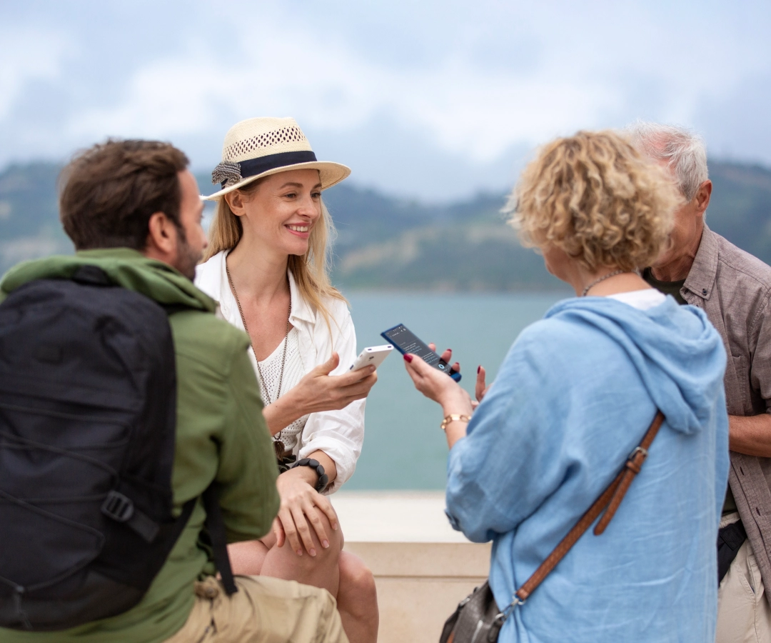 Quatre adultes, trois visibles et un partiellement obscurci, profitent d'une conversation en plein air au bord de l'eau. Deux femmes, dont l'une sourit et porte un chapeau, montrent au groupe quelque chose sur leurs traducteurs qu'elles tiennent dans leurs mains.