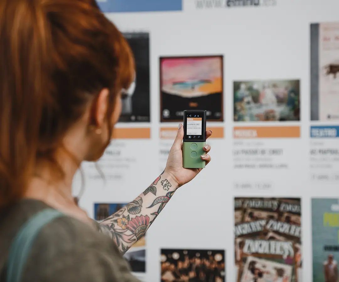 Une personne aux cheveux roux et au bras tatoué de fleurs brandit un appareil numérique vert, qu'elle dirige vers un tableau d'affichage comportant diverses affiches et informations sur l'événement.
