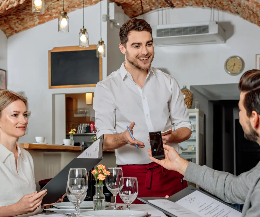 Un serveur souriant se tient à une table de restaurant, tandis qu'un client assis tient un traducteur dans sa main. Une cliente est assise à proximité et tient un menu. La table est garnie de verres, d'assiettes et d'un petit vase à fleurs.