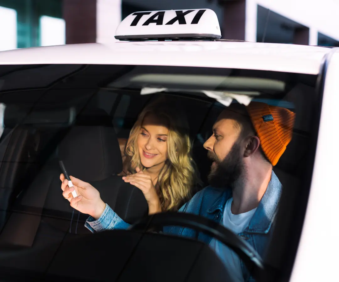 Un homme est assis sur le siège du conducteur d'un taxi et montre quelque chose à une femme sur la banquette arrière sur un traducteur qu'il tient à la main. La femme sourit avec intérêt. Un panneau de taxi est visible sur le toit de la voiture.