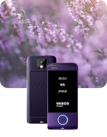 Une image de présentation du produit montrant un appareil Vasco Translator violet sous deux angles différents, sur un fond de tissu sombre et texturé. L'appareil de gauche présente une vue arrière, mettant en évidence l'objectif de l'appareil photo avec son flash et le logo « VA » en relief. L'appareil de droite présente une vue de face avec son écran allumé, affichant les messages « Hello ! », « VA », « ¡Hola ! » et le logo « VASCO Translator ». Un bouton circulaire situé au bas de la face avant de l'appareil est souligné d'une lumière bleue brillante. La composition se détache sur un arrière-plan onirique et flou de fleurs de bruyère violettes baignées d'une douce lumière du soleil.