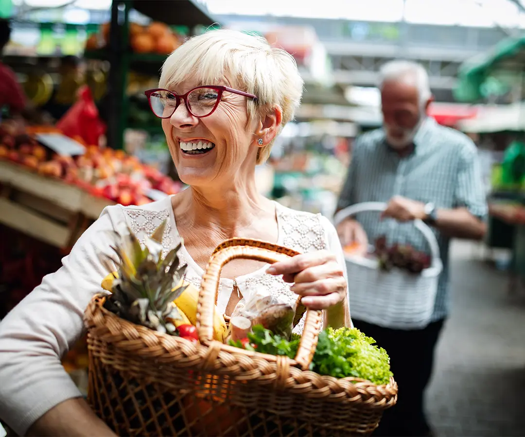 Une femme d'âge mûr, souriante, aux cheveux blonds courts et portant des lunettes rouges, tient un panier en osier rempli de fruits et légumes frais sur un marché en plein air. À l'arrière-plan, flou, une autre personne fait ses courses parmi les étals de fruits et légumes.