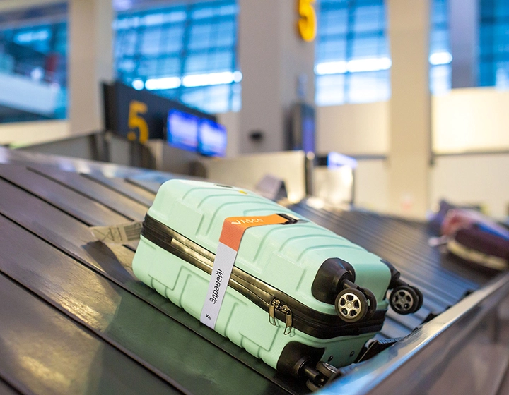 Une valise verte, sur laquelle est posée une sangle de sécurité Vasco aux couleurs de l'arc-en-ciel, repose sur le tapis roulant à bagages d'un aéroport. Grâce à cette sangle colorée, la valise est facile à repérer.